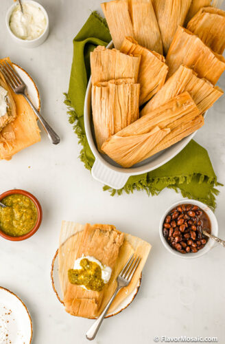 Overhead view of a white serving bowl of several tamales sitting on a green napkin with a small plate with 1 tamale topped with sour cream and green Chile salsa.