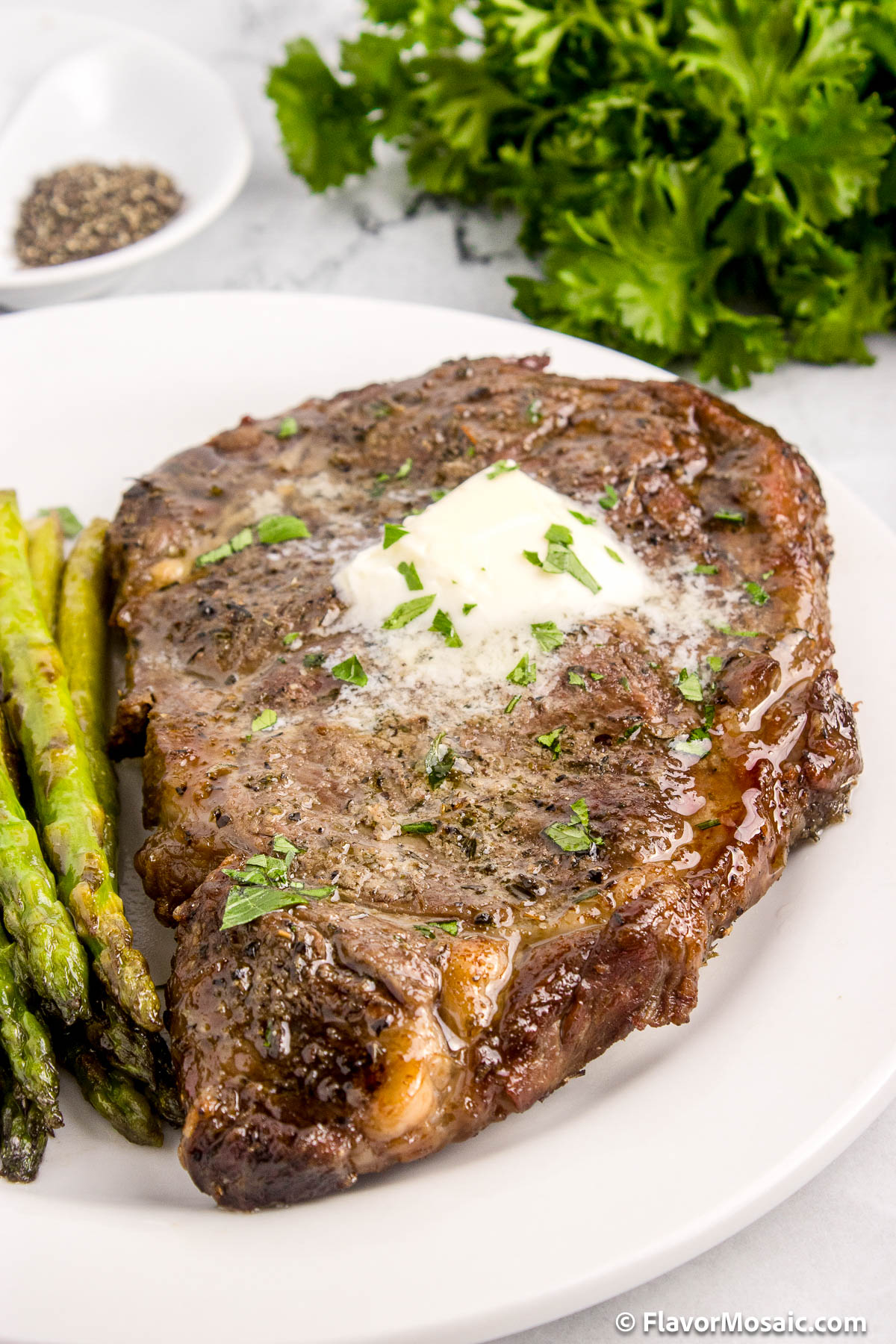 A big juicy steak (ribeye steak) with partially melted butter and chopped fresh parsley on top, sitting on a white dinner plate next to asparagus.